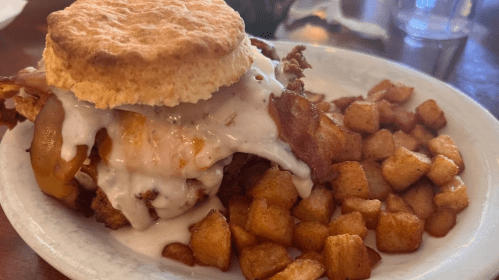 A plate featuring a biscuit sandwich with fried chicken, bacon, and gravy, served with crispy potato cubes.